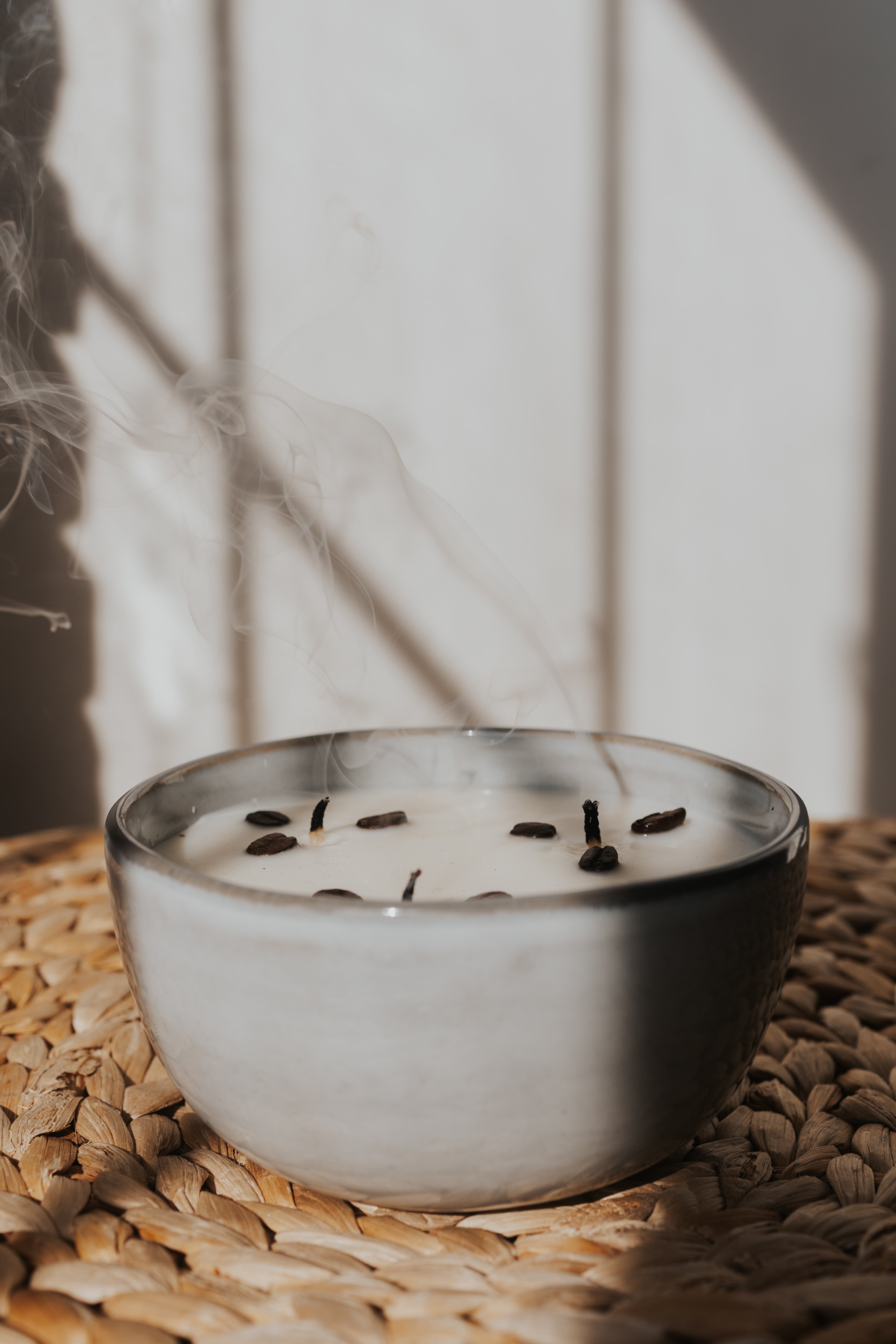 A ceramic candle in a matte gray bowl sits on a woven straw mat, with a wick and a few dark botanical pieces on top. Steam rises from the candle, and the background is a warm off-white paneled wall with soft shadows.