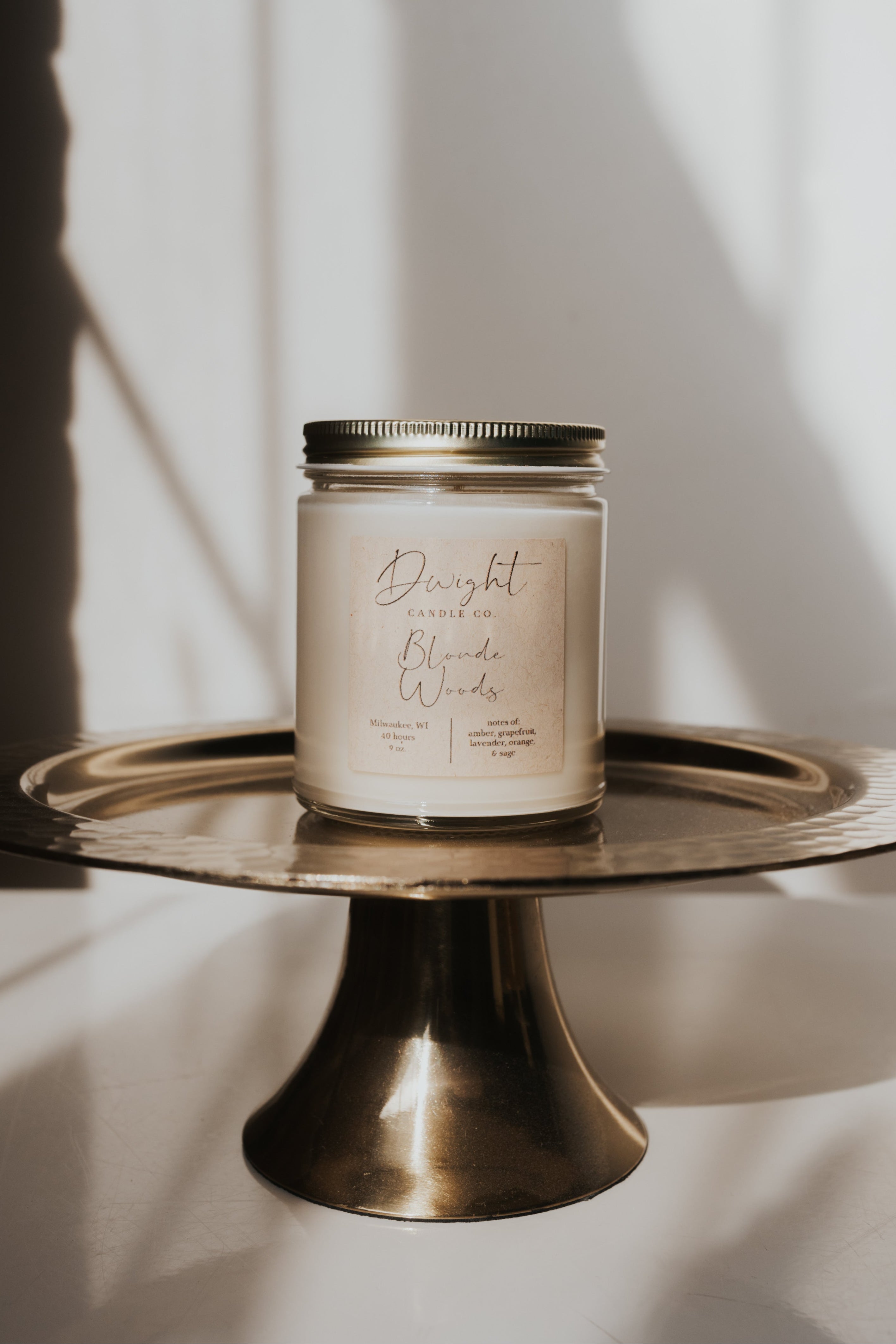 A jar candle with a cream-colored label and gold lid sits on a round brass pedestal stand against a white wall with soft window shadows.